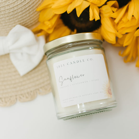 Close up of 1821 Candle Co sunflower candle with a soft floral label, styled besides a woven sun hat and fresh sunflower blooms. Ideal for cozy summer home decor inspiration.