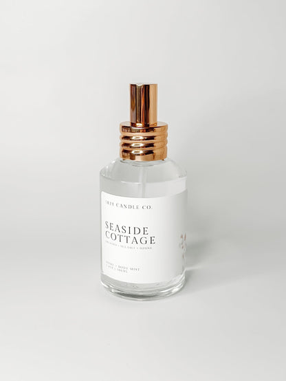 Seaside Cottage non-toxic and clean home and body mist by 1821 candle co featuring sea salt, jasmine, fresh orchid scent notes in a clear glass bottle with a gold sprayer on a white background. 