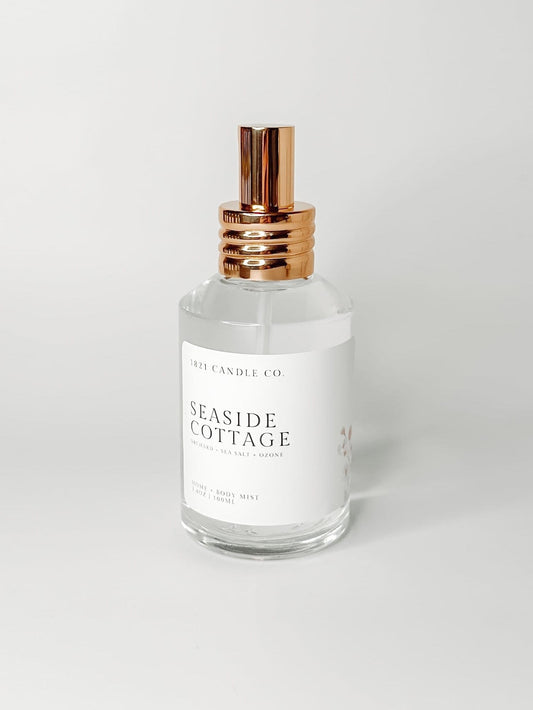 Seaside Cottage non-toxic and clean home and body mist by 1821 candle co featuring sea salt, jasmine, fresh orchid scent notes in a clear glass bottle with a gold sprayer on a white background. 